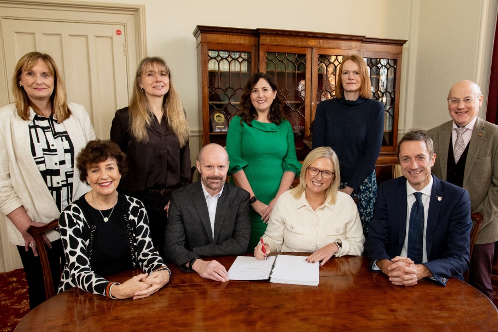 Seated in the Hamilton room with Queen's and Belfast Trust representatives standing and sitting, Gillian Magee signs Belfast Trust contract extension